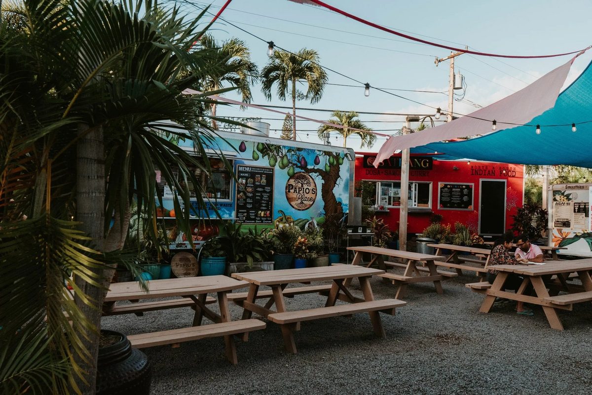 Food trucks and picnic tables at the South Maui Gardens truck park in Kīhei, with a couple sharing a meal under the shade sails